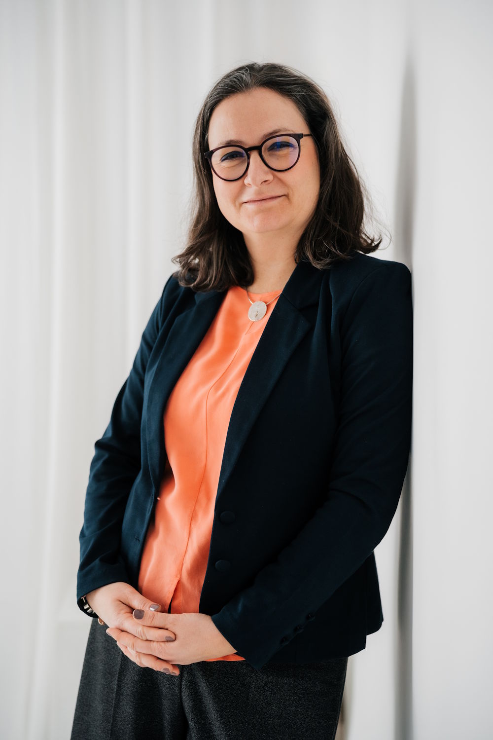 Coach für mehr Balance in Beruf und Beziehungen lehnt entspannt an einer Wand und trägt dunkles Outfit mit orangefarbener Bluse